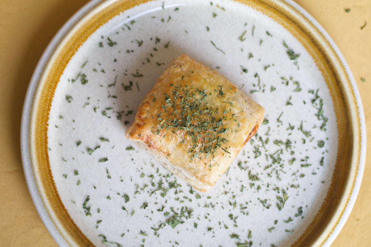 A baked square pastry or puff pastry with a golden-brown crust, topped with chopped green herbs, served on a white ceramic plate with a gold rim on a yellow surface.